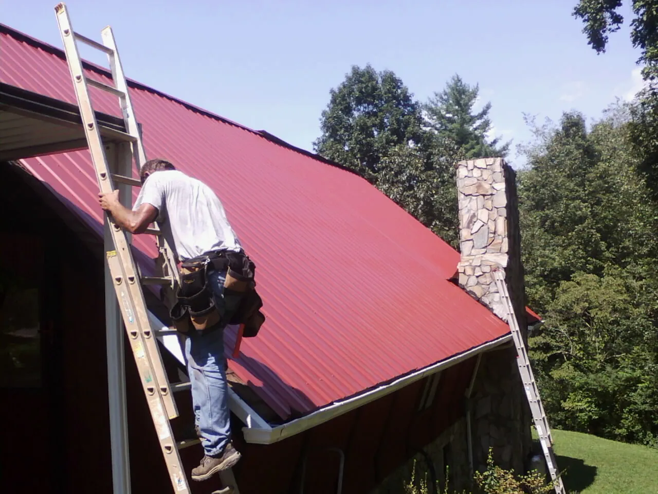 Roofer on a ladder beside a red metal roof and stone chimney — Soffit & Fascia Repair by Roofing Brothers in Lake Jackson